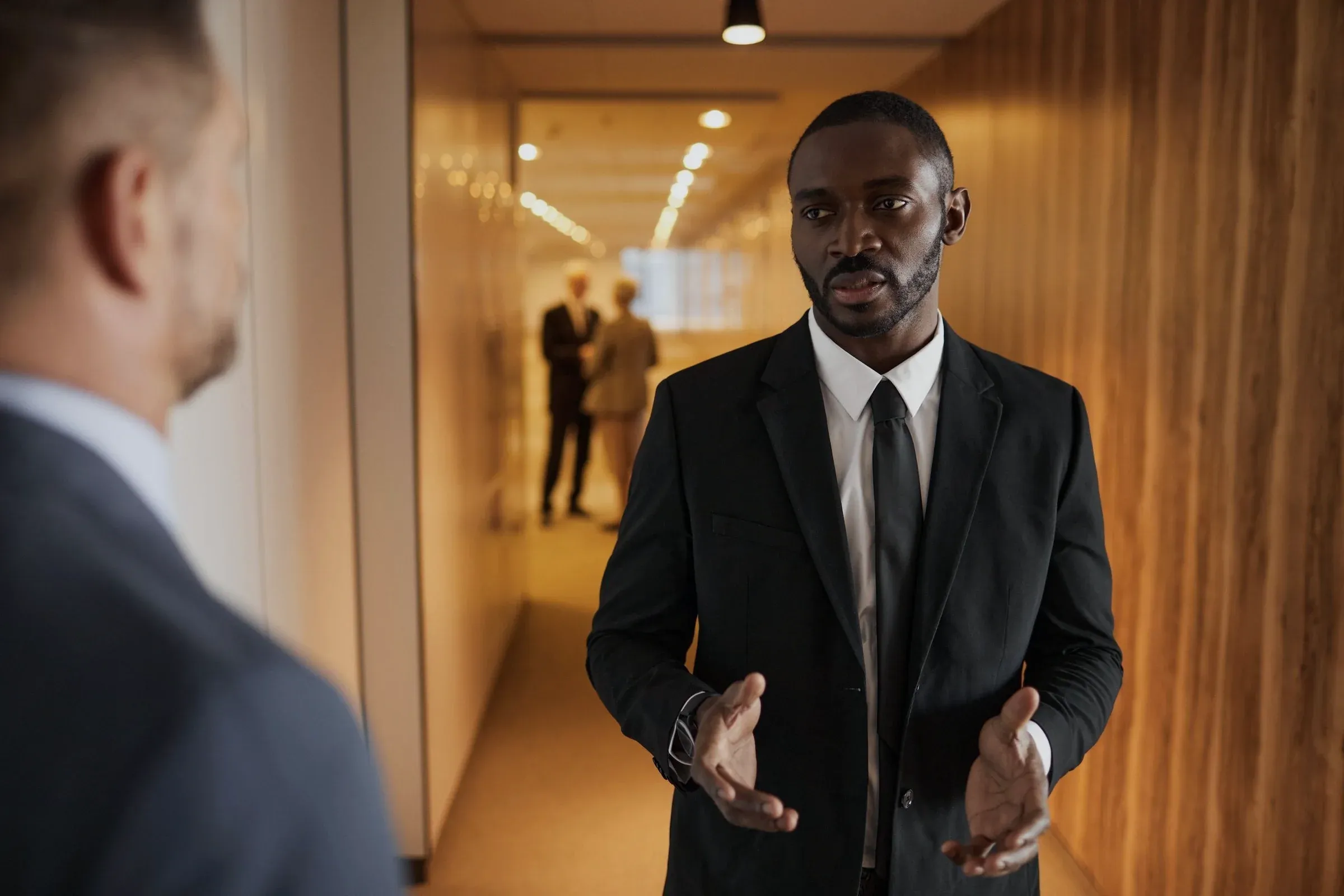 An executive in a dark suit talking with a colleague in a glass-walled corridor
